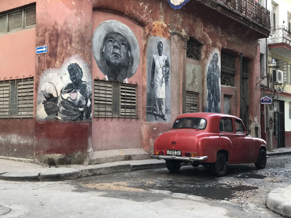 Old red car in front of a a wall with murals in Cuba